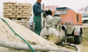 Budowlaniec wsypuje cement do miksokreta. Obok stos worków z cementem i góra piasku.