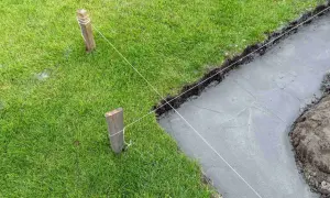 Wyznaczanie fundamentów domu za pomocą sznurka i palików. Widać świeży beton i trawę.