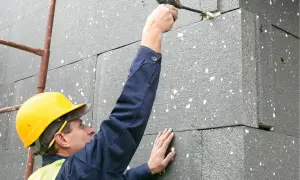 Budowlaniec w żółtym kasku i kamizelce nakłada piankę montażową na styropian. Grubość styropianu jest kluczowa dla izolacji.