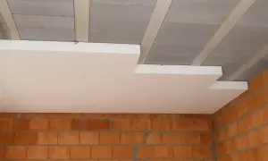 Białe płyty styropapa montowane pod betonowym stropem, nad ceglanymi ścianami. Izolacja termiczna w trakcie budowy.