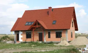 Dom w stanie surowym otwartym, z czerwonym dachem i oknami dachowymi. Wokół materiały budowlane i piasek.