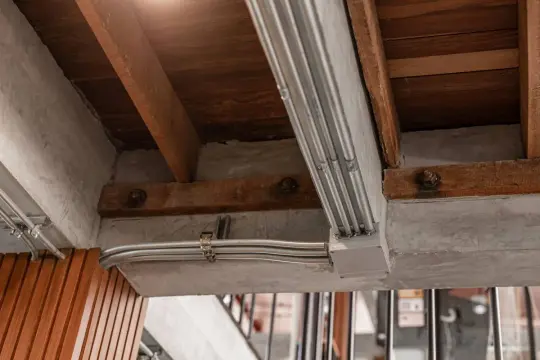 Metalowe rury elektryczne biegną wzdłuż betonowego podciągu, łącząc się w puszce. Drewniane belki stropowe i beton tworzą surową, industrialną estetykę.