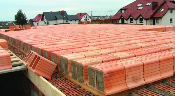 Strop ackermana w budowie, rzędy pustaków ceramicznych czekają na zalanie betonem. W tle domy z czerwonymi dachami.