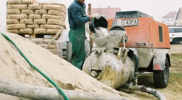 Budowlaniec wsypuje cement do miksokreta. Obok stos worków z cementem i góra piasku.