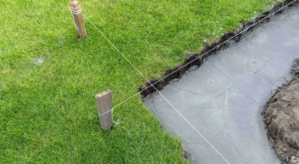 Wyznaczanie fundamentów domu za pomocą sznurka i palików. Widać świeży beton i trawę.