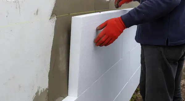 Ręce w czerwonych rękawiczkach układają płyty styropianu na ścianie. To etap budowy domu ze styropianu.