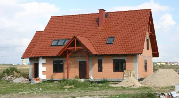 Dom w stanie surowym otwartym, z czerwonym dachem i oknami dachowymi. Wokół materiały budowlane i piasek.