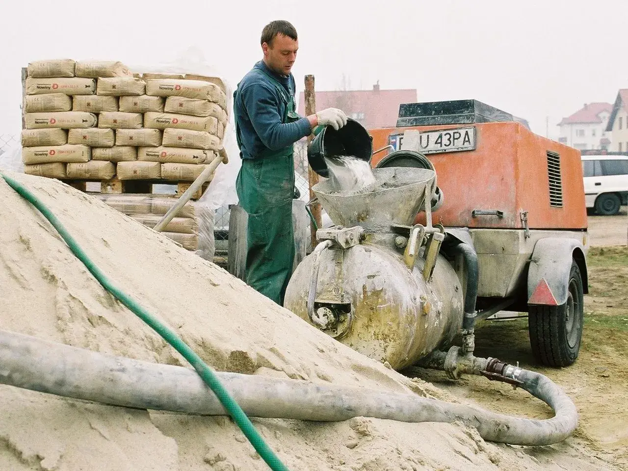 Budowlaniec wsypuje cement do miksokreta. Obok stos worków z cementem i góra piasku.