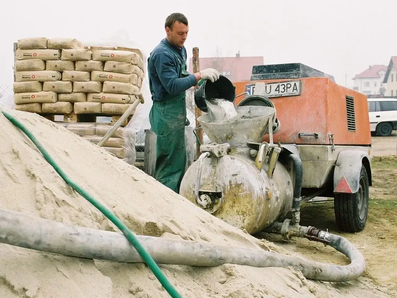Budowlaniec wsypuje cement do miksokreta. Obok stos worków z cementem i góra piasku.