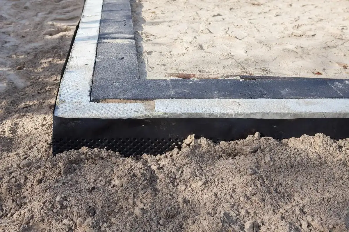 Warstwa izolacji poziomej fundamentów chroni przed wilgocią. Widoczna na piasku, z czarną membraną i warstwami betonu.