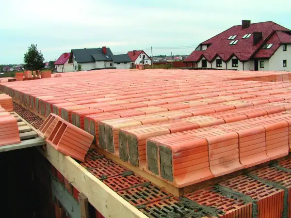 Strop ackermana w budowie, rzędy pustaków ceramicznych czekają na zalanie betonem. W tle domy z czerwonymi dachami.