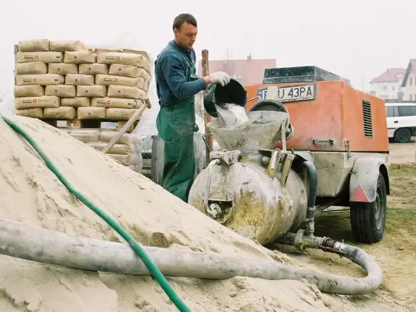 Budowlaniec wsypuje cement do miksokreta. Obok stos worków z cementem i góra piasku.