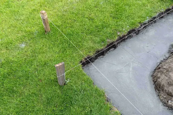 Wyznaczanie fundamentów domu za pomocą sznurka i palików. Widać świeży beton i trawę.