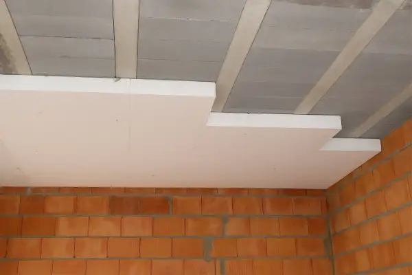 Białe płyty styropapa montowane pod betonowym stropem, nad ceglanymi ścianami. Izolacja termiczna w trakcie budowy.