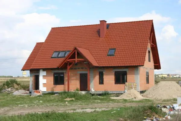 Dom w stanie surowym otwartym, z czerwonym dachem i oknami dachowymi. Wokół materiały budowlane i piasek.