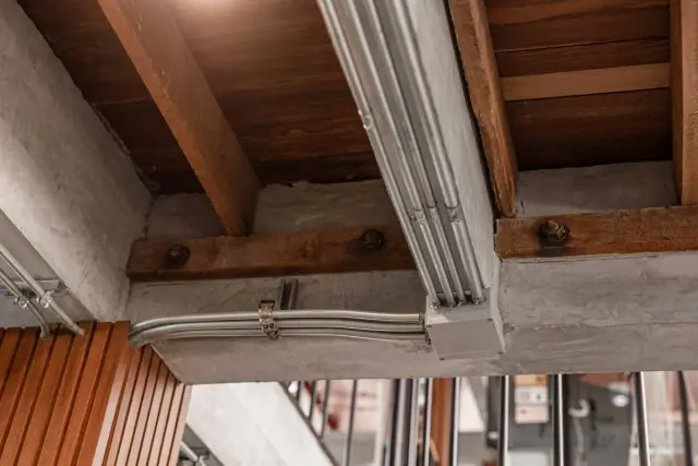 Metalowe rury elektryczne biegną wzdłuż betonowego podciągu, łącząc się w puszce. Drewniane belki stropowe i beton tworzą surową, industrialną estetykę.