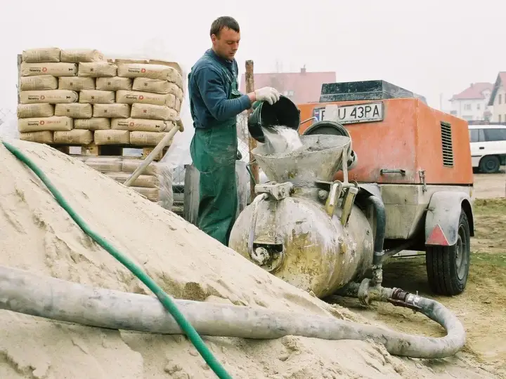 Budowlaniec wsypuje cement do miksokreta. Obok stos worków z cementem i góra piasku.